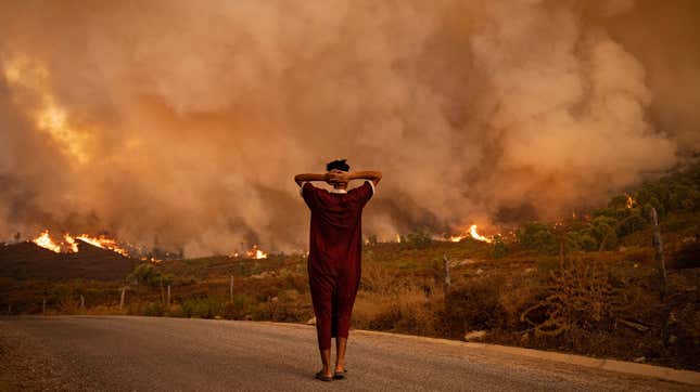  woman looks at wildfires tearing through a forest in the region of Chefchaouen in northern Morocco on August 15, 2021. Smoke and flames rise in the background as she clasps her hands behind her head.