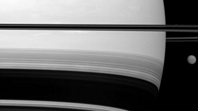 A black and white image of Titan passing to the side of Saturn.