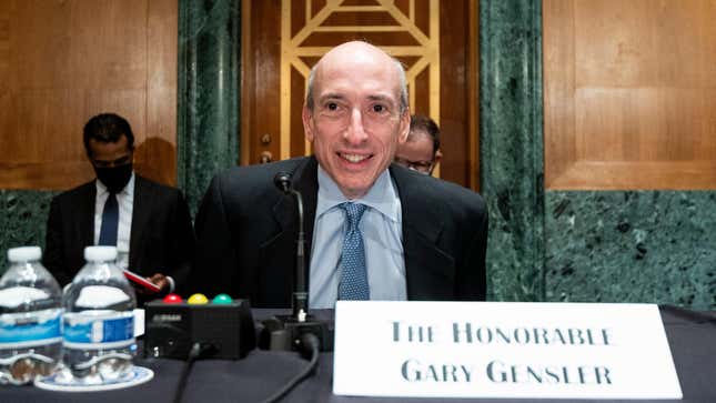 A balding man sits behind a desk with a microphone in front of him. A placard on the table reads "The Honorable Gary Gensler."
