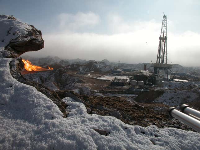 Natural gas burns from an oil well where a large metal structure rises over a leveled-out ground. Snowy mountains are in the distance.