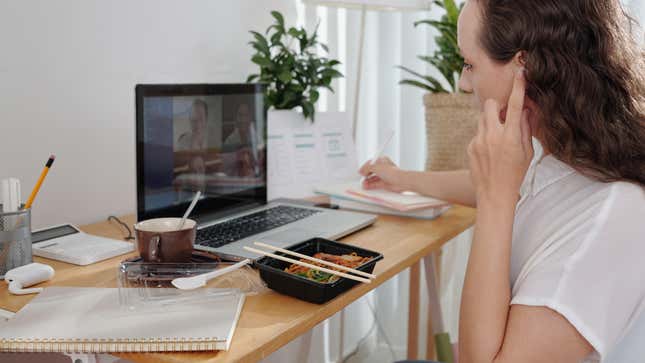 A woman works at a laptop while eating lunch and taking notes in a notebook