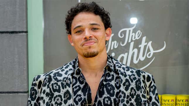 Anthony Ramos, dressed in a silver and black faux leopard print suit, smiles in front of an In the Heights promo board.