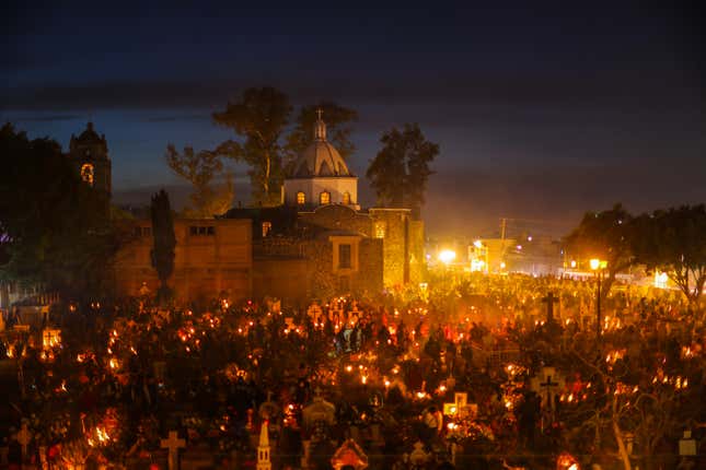 A church is seen in the background, illuminated by the glow of street lights and candles being held by people in a large crowd.