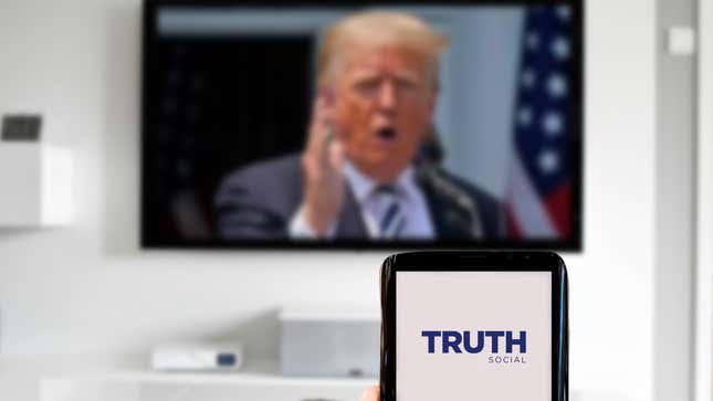 Truth Social logo displayed on a smartphone against blurred background with Trump on TV