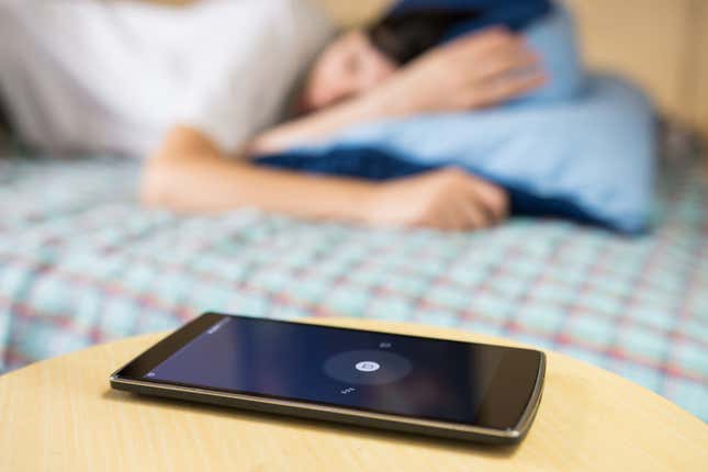 A smartphone sits on a bedside table with the screen illuminated showing an alarm, with a woman sleeping out of focus in the background