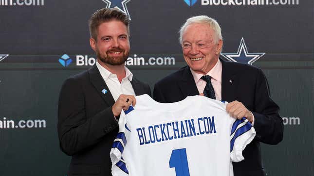 Blockchain.com CEO Peter Smith holds a jersey emblazoned with the number 1 and "Blockchain.com" alongside Dallas Cowboys owner Jerry Jones, both smiling.