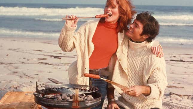 1970s photo of cuddling couple grilling hot dogs on the beach