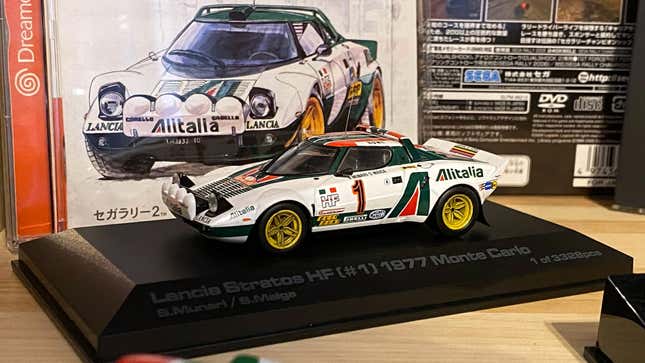 Photo of a 1/43-scale Lancia Stratos model car in front of an illustration of the Stratos from the Sega Rally 2 Dreamcast game box.