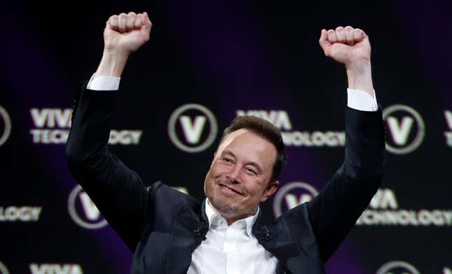 Elon Musk in a black suit with a white button up shirt holding his fists up in victory with a smirk on his face. The background is a blackdrop with a white logo which reads VIVA TECHNOLOGY