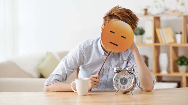 A man sits slumped at a table with a coffee cup and alarm clock on it, his face covered by a lazy emoji