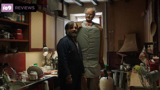 A man in a blue jacket and a very tall man-robot wearing a bow tie stand in a cluttered kitchen in a scene from Brian and Charles.