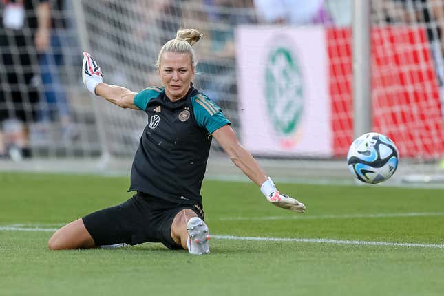 A white women with blonde hair in a ponytail does the splits in attempting to reach a soccer ball headed into the net.