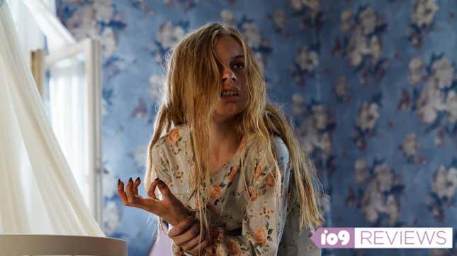 A 12-year-old girl with long blonde hair holds her wrist and snarls in a room covered in blue wallpaper.