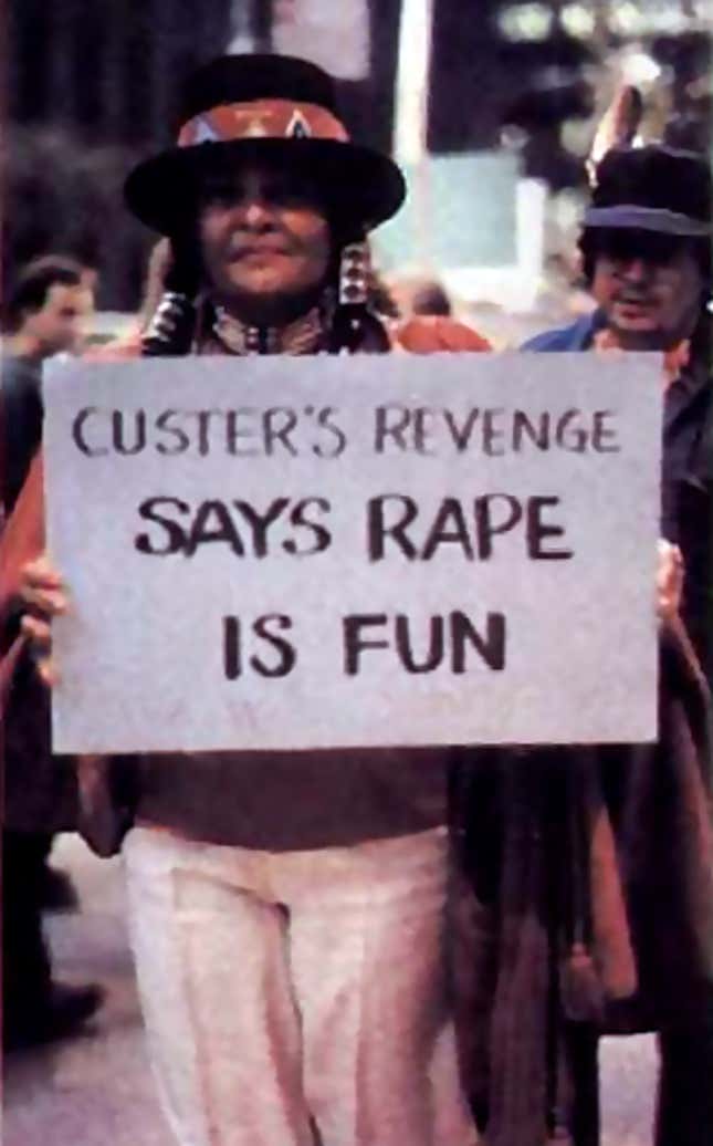 A woman wearing Native American signifiers holds a protest sign that says "Custer's Revenge says rape is fun".
