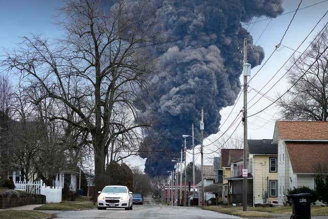 A view of a suburban street lined with houses and bare trees. In the distance rises a colossal column of dark smoke set against a blue sky. 