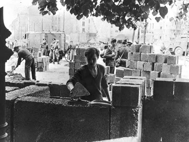 A worker builds the wall, watched by East German soldiers