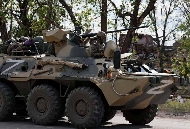 A camouflaged tank with a Z painted on it traveling on a rode with a soldier sitting on it holding a rifle.