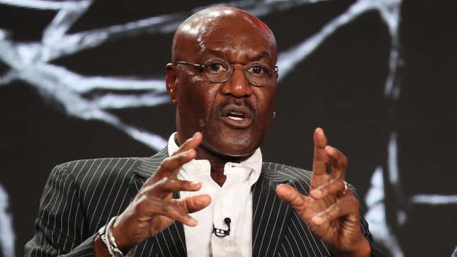 Delroy Lindo speaks at an event with his hands raised. He's wearing a mostly black striped suit jacket, white shirt, and glasses.