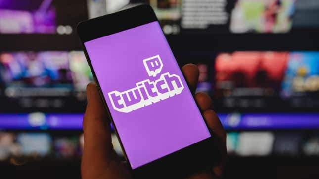 A hand holding a phone with the Twitch logo in front of a screen displaying multiple twitch channels.