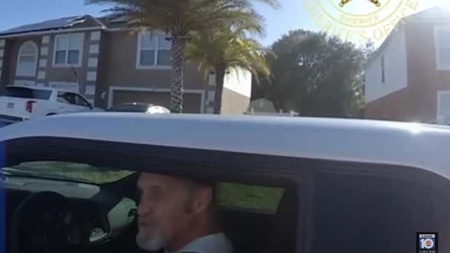 A man with a gray beard sits in a stopped white car on the shoulder. There are palm trees and ugly, cheap Florida houses in the background
