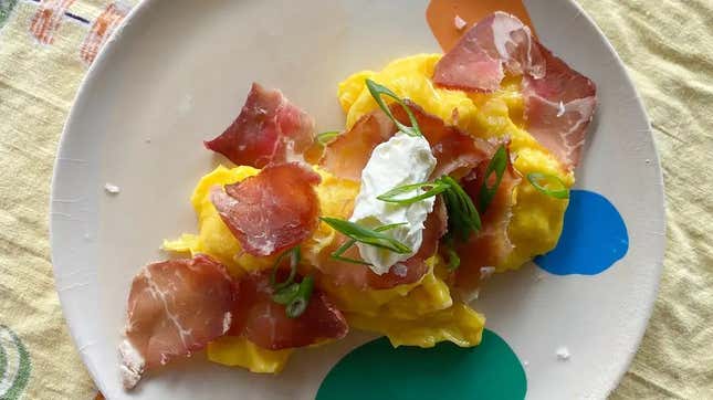 A serving of scrambled eggs on a plate, topped with thinly sliced meat and a dollop of sour cream and chives