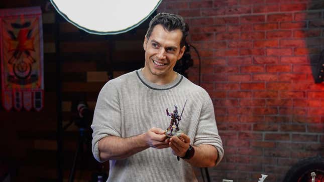 Henry Cavill smiles while holding a Warhammer miniature as he hears the Monkey's Pawl tighten. 