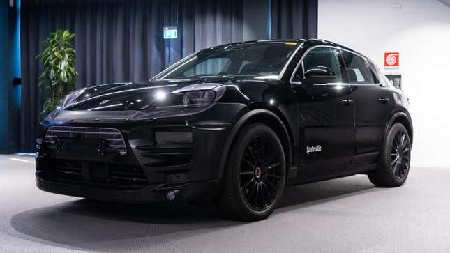 A 2024 Porsche Macan Electric prototype photographed inside a demonstration hall. the vehicle is painted black with vinyl appliques to obscure its design