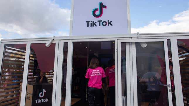 A large glass panneled building with the Tiktok logo on a sign above a door. A person stands in from of the door with a shirt that says "ask me anything about TikTok.
