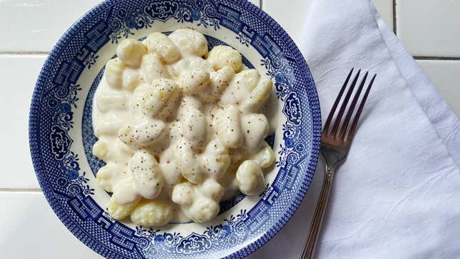 A bowl of gnocchi on a table in a blue china dish, the pasta covered in a creamy white sauce flexed with specks of black pepper