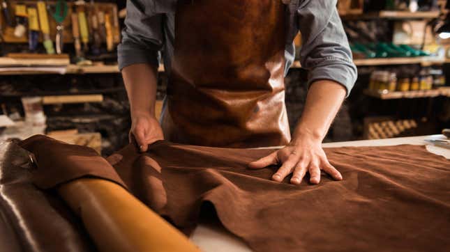 A man lays out a piece of leather on a worktable in a shop with leather working tools visible hanging on the wall behind him
