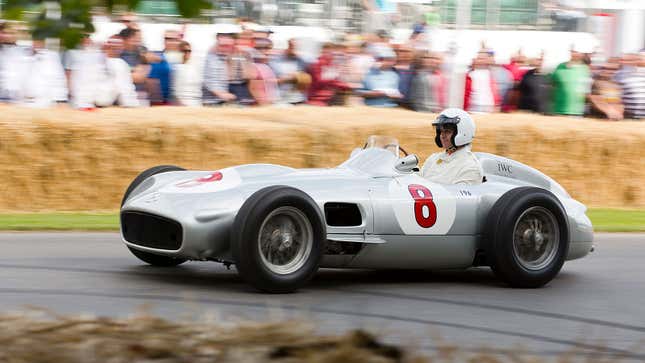 A photo of a silver 1954 Mercedes F1 car racing up a hill. 