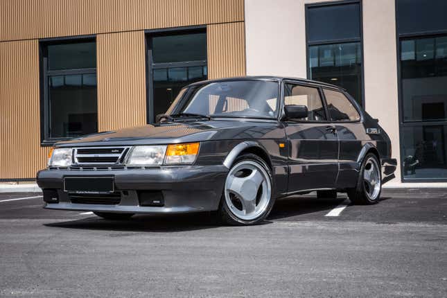 A grey 1980s Saab 900 Tubo on three-spoke wheels is parked in front of a building.
