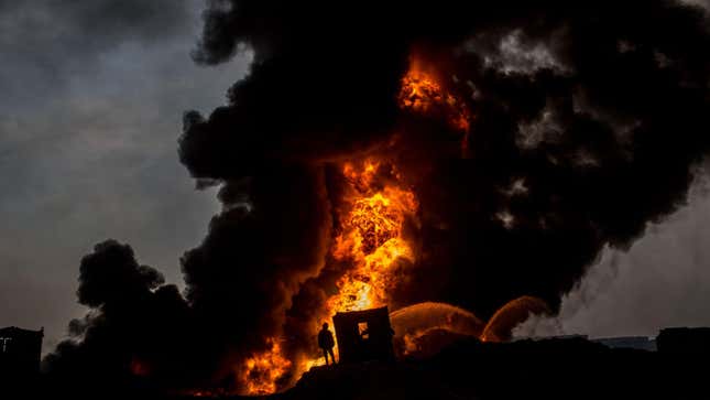 A firefighter works to extinguish an oil well set on fire as plumes of smoke rise against a dark sky.