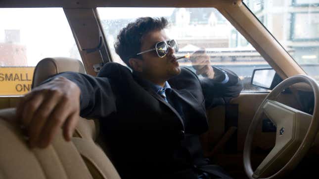 A man wearing a suit and dark sunglasses in the driver's seat of a vehicle lights a smoke.
