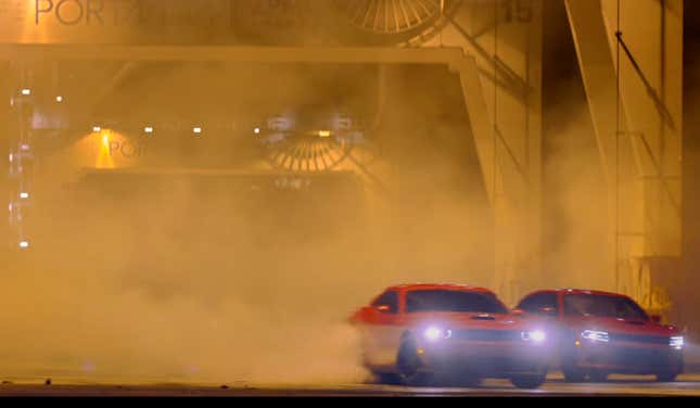 Two red cars--a Dodge Challenger Hellcat and a Dodge Charger Hellcat--drifting and kicking up  smoke from their tires in front of a port entrance. 