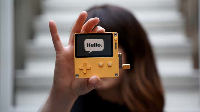 A person holds up a small, yellow electronic device with the word "Hello" on its screen.