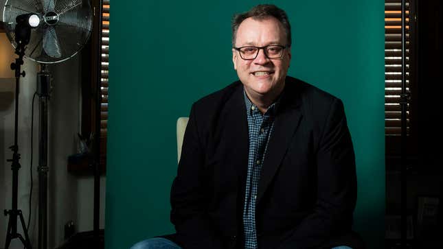 Russell T. Davies poses in a new photo released by the BBC to accompany the announcement of his return to Doctor Who in 2023. He's sitting, wearing a black suit jacket over a blue button down shirt and in front of a green paper background.