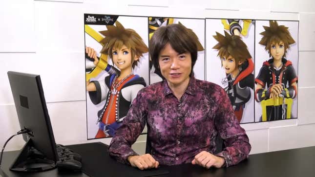 Masahiro Sakurai sits at a desk, wearing a purple shirt. 