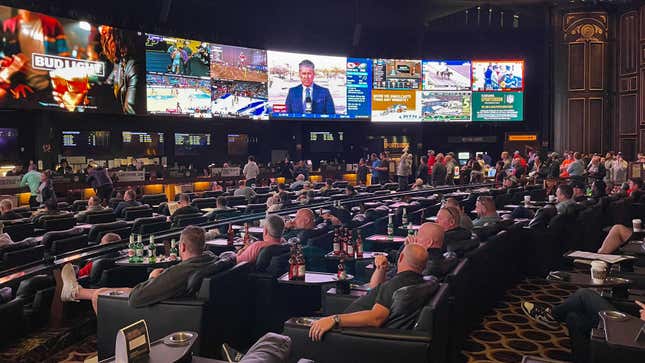 A large room with multiple large-screen TVs in a ring around the room show various NFL games and betting lines. Dozens of middle-aged men sit in recliners in front of the screens, many with beer bottles on tables in front of them.