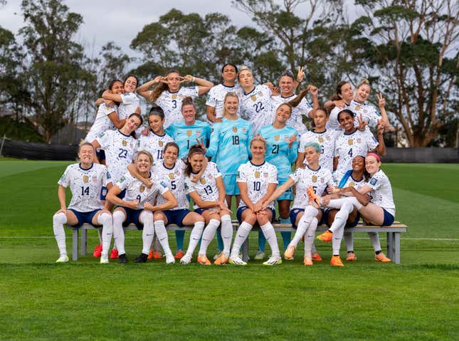 A group of women in matching white jerseys and blue shorts smile and mug for the camera in an outdoor team photo.