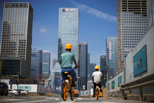 Two men in yellow hardhats ride bicycles on the road while skyscrapers, including the Evergrande headquarters, rise in the distance into a blue sky.