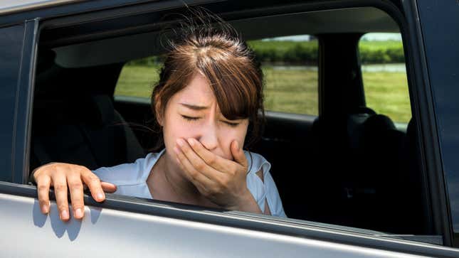 A Young brown-haired woman leans her head out of a car window while holding a hand to her mouth with a pained expression
