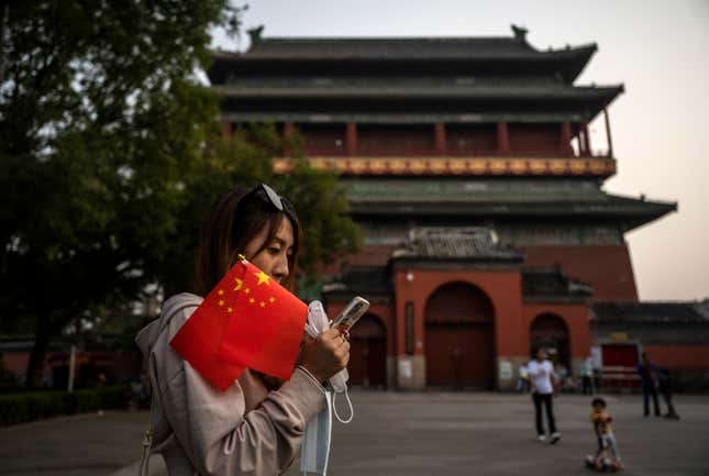 A woman holds two Chinese flags in her hand while looking at her mobile phone. The Beijing Drum Tower is visible in the background.