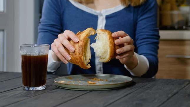 Hands tearing croissant in half beside cup of coffee