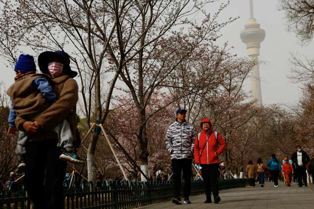 People walk through a park lined with budding trees. The sky is hazy from a sandstorm. The Beijing TV Tower rises in the distance.