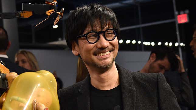 A middle-aged Japanese man in thick, circular glasses smiles at something off-camera.