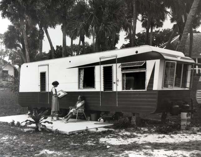 A woman with a clipboard interviews an individual who is seated outside of a mobile home.