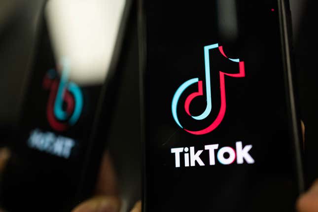 A black smartphone screen shows the TikTok logo. The phone is next to a surface that shows its blurry reflection.