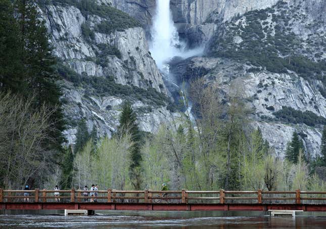 People cross a bridge over the Merced River, which is swollen almost to the bridge level, with Yosemite Falls in the background. 