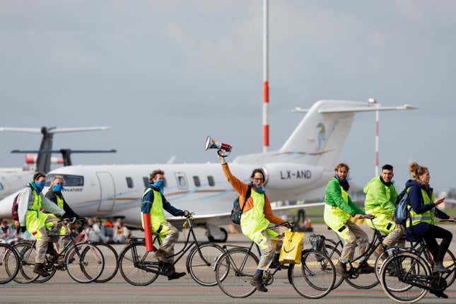A group of protestors in neon yellow aprons ride bicycles in front of a private jet on the tarmac. One cyclist is raising an arm holding a megaphone.
